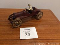 Front-side view of antique cast iron toy car with removable driver seated inside, placed on wooden surface with auction tag.