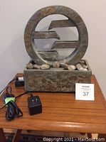 Front view of the Sunnydaze stone circular water fountain placed on a wooden surface, showing the circular design with horizontal stone bars, pebbles, pump cord, transformer, and auction tag.