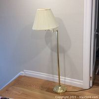 Full view of brass finish standing lamp with shade shown against wall and hardwood floor.