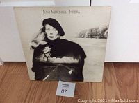 Front cover of the Joni Mitchell Hejira vinyl album showing the artist portrait with a beret and a cigarette, background is a blurred road and trees, on wooden floor.