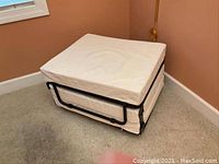 Compact folded position of the cot, showing it doubled as a square hassock or ottoman with cushioned top and a black metal frame visible on sides.