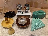 Photo of Art Deco style kitchenware and decorative items on a counter showing metal creamer and sugar with orange handles, ceramic pieces in turquoise and yellow, brown bowl, silver bowl, and black ashtray.