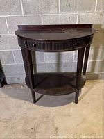 Front view of vintage half round entry table against concrete wall, showing the single drawer and bottom shelf.