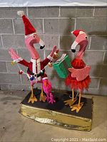 Front view showing two pink flamingos in Santa hats and holiday attire on gold base with decorations and lights.