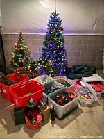 Two artificial lighted Christmas trees, one small and one large, standing with a lighted wreath and multiple bins containing holiday lights and decorations.