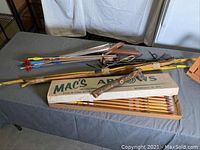 Wide shot showing bows, arrows, quiver, bowstrings, and Mac's Arrows box on a table.