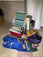 Full view of camping items including blue collapsible cot in bag, cooler, fishing rods, camping accessories, and folding chair.
