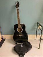 Front-facing full shot of the black Gibson Maestro acoustic guitar standing on a stand with a black gig bag case on the floor in front.