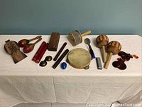 Photo showing entire lot of small percussion instruments arranged on a white tablecloth with a blue-gray wall background.