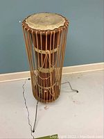 Full side view of wooden Bongo Congo drum with taut animal skin top, rope tension system, and metal tripod stand on floor.