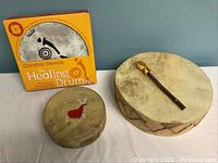 Healing Drum Kit box alongside two hand drums and a wooden mallet laid out on a white surface against a blue wall.