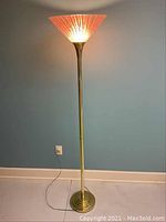 Full view of the lit floor lamp against a blue wall and white baseboard showing the orange art glass shade glowing.