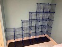 Blue modular wire rack assembled against room corner showing bent and intact grid panels creating several open cubical compartments, some bent panels visible on bottom left side.