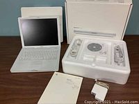 Apple iBook G4 laptop open and powered off, showing white keyboard and screen, placed next to original box with power adapter, software CD, cables, and manual