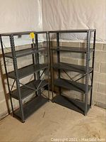 Two metal shelving units standing side by side in a basement against a concrete block wall with plastic sheeting above. Each has five shelves and perforated upright posts with hole pattern for shelf adjustment.