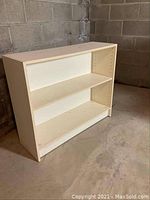 Front angled view of white low bookshelf showing two shelves and pre-drilled holes on the sides for shelf adjustment.