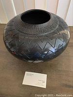 Front view of black pottery vessel with black chevron pattern and textured neck, shown with a card indicating details.