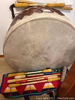 Front view of pow wow drum leaning against wall with two sticks on top and eight sticks plus smudge bundle on patterned blanket