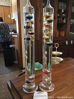 Photo showing both glass Galileo thermometer barometers standing side by side on a wooden table with background furniture and a person in the background, showcasing the full height and colored floating bulbs inside.