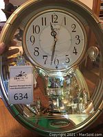Front view of the musical clock showing white dial with black Arabic numerals, silver-tone frame and Rhythm brand badge.
