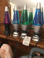 Photo of six lava lamps with silver bases and caps in colors including purple, blue, and green, cords wrapped around bases, showing tops and bottoms