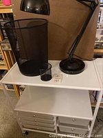 White rolling craft table with black mesh trash can, small pencil holder, and black desk lamp on top