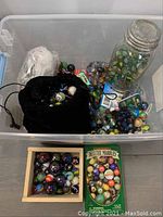 Clear plastic bin holding a metal tin, wooden box, glass jar, velvet pouch and loose marbles