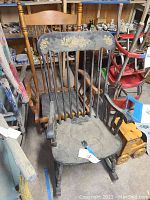 Two wooden rocking chairs together. One chair has a dark painted floral design on the top rail, the other chair is stained natural wood. Visible wear and dust, set indoors amongst other items.