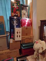 Stack of nine assorted boxes of Christmas decorations and figurines including a boxed Santa doll, Christmas tree light and wrapped fabric item.