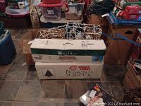 Boxed 6 ft artificial Christmas trees stacked with outdoor reindeer decoration on top and storage bins in background