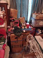 Full view of stacked boxes and assorted Christmas decor items in a room corner, including green string lights and tinsel garland.