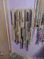 Image of a cork board mounted on a purple wall with various necklaces hanging, showing different styles including faux pearls, chains, and beaded strands.
