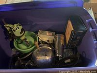 Top-down view of various vintage kitchen appliances and tools inside a blue plastic container including a mixer, strainer, grinder, and shaker cup.