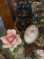 Photo shows iridescent green and purple glass pedestal bowls, porcelain rose figurine, and porcelain oval box decorated with butterflies and flowers.