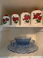 Four white ceramic canisters with red rose designs arranged on a shelf above blue glass serving items.