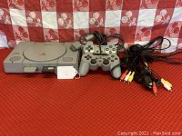 Sony PlayStation original console with two wired controllers and cables on red patterned surface.