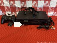 Front view of Sony PlayStation 2 console with label tag, two wired controllers, and AV cable laid out on red mat with patterned cloth background.