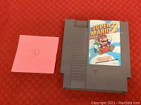 Top view of the Nintendo NES Super Mario Bros 2 grey game cartridge with label shown frontally, resting on a red checkered surface with a pink sticky note numbered 30.