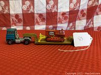 Side view of blue and silver Matchbox Super Kings low loader truck carrying an orange bulldozer on yellow flatbed trailer on red patterned fabric background.