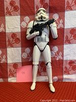 Front-facing image of 13-inch Stormtrooper action figure holding a black blaster rifle on a red and white patterned background.