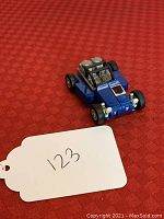 Blue dune buggy toy with black roll cage and detailed engine, shown on a red textured surface with a white tag labeled '123'