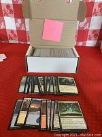 White storage box open, showing approximately 400 loose Magic the Gathering cards inside. White and black bordered cards are separated. A small number of cards are spread loose in front of the box showing multiple card types and colors.
