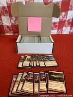 White cardboard box open showing approximately 400 Magic the Gathering cards stored vertically with some cards laid out in front showing artwork of red themed cards and creature spells.