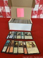 Closed white cardboard box filled with stacked Magic the Gathering black border cards, with a pink note '138' on the inside lid and a sample of loose cards laid out in front showing varied artwork and card types.