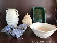 Front view of the five ceramic pottery pieces arranged on a wooden shelf: white ribbed bonsai planter with loop handles, cream covered jar with lid, cream-white ribbed vase, black ruffled-edge bowl, and cream-white bowl.