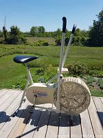 Side view of the Vitamaster elliptical exercise bike on wood decking outdoors, showing the seat, handles, pedals, and fan wheel.
