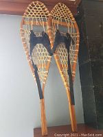 Pair of wooden snowshoes shown leaning upright with black bindings visible on both. Wooden frame and woven netting detail clear.