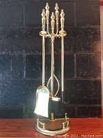 Front view of the brass fireplace tool set with poker, shovel, broom, tongs, and stand against black background.