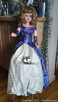 Full view of the Deluxe Reading Sweet Ann doll in floor-length purple satin gown with white netted underskirt, standing on wooden floor in front of kitchen cabinets