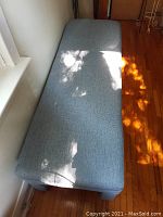 Overhead photo showing the rectangular top of the upholstered bench covered in blue linen-look fabric with some light reflections.